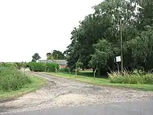 Small farm track leading off a mintor road. behyind a low hedge some single story brick farm buildings peep. A huge tree ominates the right side of the picture