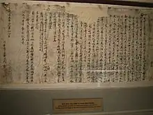 A tattered, ripped and crumpled piece of light coloured paper with writings penned in Chinese chearacters in dark ink, pinned on a wall for display in a museum.