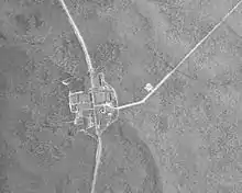 Black and white aerial view of a forested area with a vertical curving highway running through a group of large buildings near the center. A smaller road runs diagonally to the top right corner.