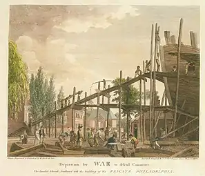 Carpenters work in the foreground, while the wood is hauled up a scaffold to the almost-complete hull.