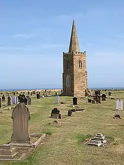 A two-stage church tower, topped with a steeple, surrounded by gravestones, and a view of the sea beyond