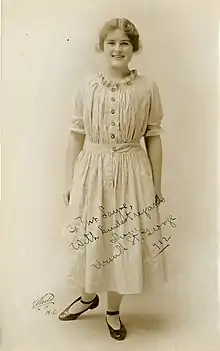 A young smiling white woman, standing, wearing a light-colored dress with buttons down the front of the bodice and a ruffled neckline.