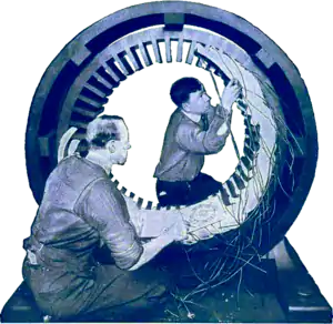 Blue-tinted photograph of two men working on a large circular piece of electrical equipment.