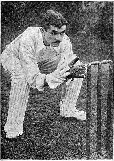 Man with moustache in wicket-keeping position, crouching behind stumps.